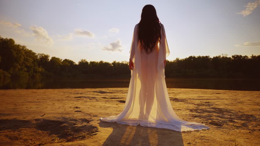 Happy woman walks along beach river banks on sand enters the water. Girl back rear view enjoy nature free green tree magic sun light sky. Lady walks in silk white long dress fly in wind slow motion. Royalty-Free Stock Footage #1106747493
