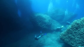  scuba divers exploring around a reef underwater deep blue water big rocks and bubbles ocean scenery 