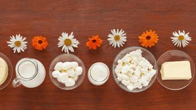 Summer fresh natural dairy products are arranged in row on wooden table. Organic dairy food - milk, curd, cheese, Greek yogurt, mozzarella, butter and chamomile, marigold flowers. Flat lay, video