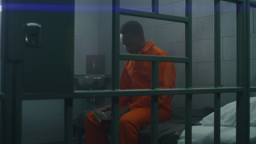 African American prisoner in orange uniform sits on the bed behind bars, reads Bible in prison cell. Criminal serves imprisonment term for crime in jail. Detention center or correctional facility. Royalty-Free Stock Footage #1106769625