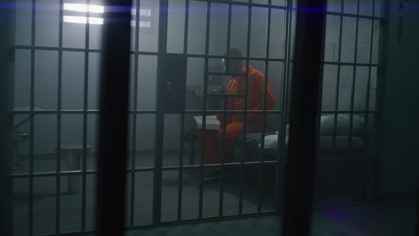 African American prisoner in orange uniform sits on the bed, reads Bible in prison cell. Prison officer walks, watches criminal. Inmate serves imprisonment term for crime in jail or detention center.