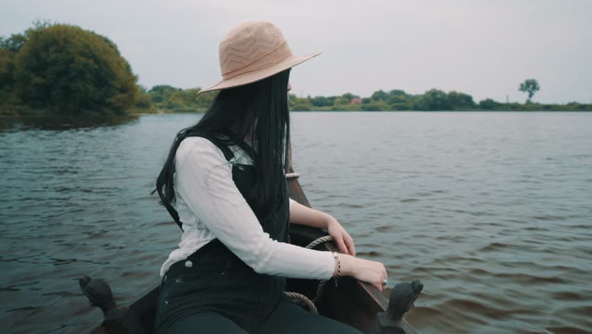 A girl in a hat sits in a boat floating on a river and looks at the landscape
