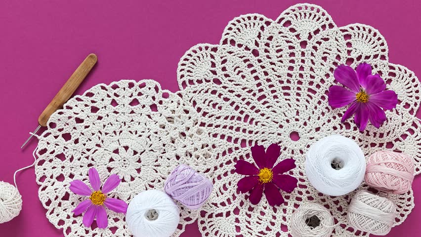 Video of summer needlework and craft. Two round handmade lace napkins, skeins of cotton yarn, crochet hooks and cosmea flowers on pink background. DIY concept. Top view, close up, flat lay
