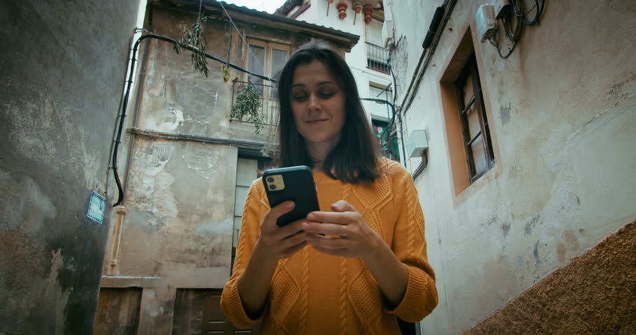Attractive tourist woman walking through old city street on vacation and using smartphone for navigation or communication with friends