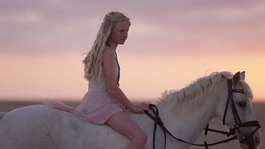 A medium shot of a blonde girl in a white dress bareback riding a white horse against the orange sunset