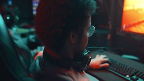 Close-up rear shot of unrecognizable young man in glasses, with curly hair sitting in cyber cafe, preparing to play shooter video game and putting on headset