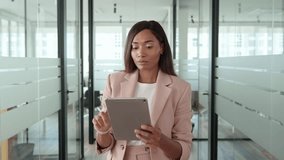 Young busy professional African American business woman company manager sales executive wearing suit holding tab, using digital tablet computer standing in office looking at pad thinking.
