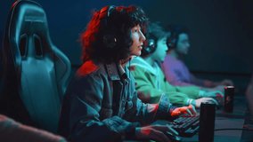 Medium shot of young biracial girl with curly hair, in headphones sitting at computer in cyberclub, playing video game, failing, leaning back on chair and sipping energy drink from can