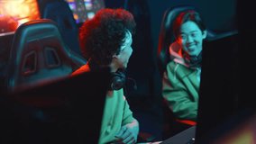 Medium shot of two cheerful young multiracial and Chinese men chatting and laughing in cyber club, then putting on headphones and beginning to play FPS video game