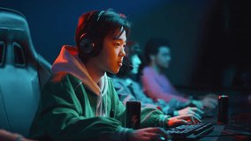 Medium side shot of young Chinese man in headphones sitting at computer in cyberclub, playing video game with diverse friends and sipping energy drink from can
