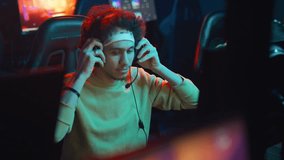 Medium shot of young multiethnic guy with curly hair, headband and headset sitting in cyberclub at night, playing shooter video game and sipping energy drink from can