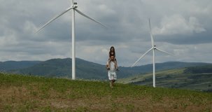 This video captures a heartwarming scene of a happy family, with the father and his daughter strolling through a field, with the little girl sitting on her father's shoulders, and wind turbines wind