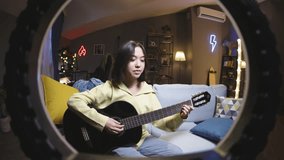 Asian girl blogger plays guitar and sings. Talented creative girl is trying to record video of playing guitar, while her stupid silly restless friends spoil frame and video, writhing and grimacing.