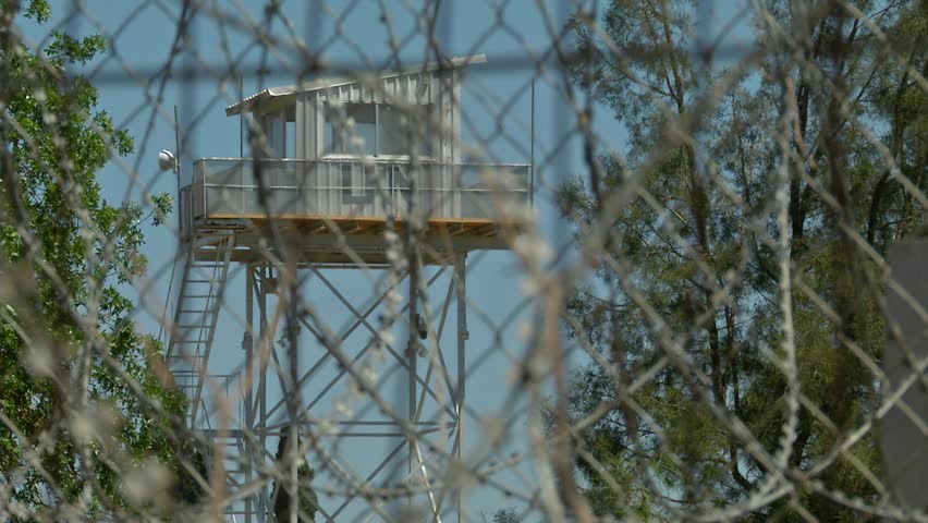 A tall, metallic watchtower is visible behind barbed wires in a sunny day. the camera is changing focus towards the sharp fence and back to the tower. some heat haze is apparent also. Nicosia, Cyprus.