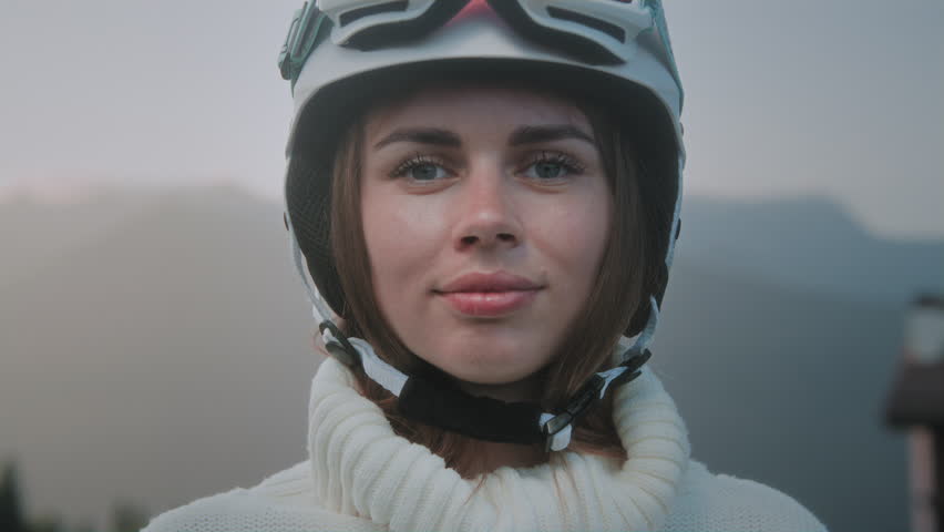 A beautiful Caucasian girl puts a white ski or snowboard helmet on her head. Portrait of a pretty woman at a winter resort. The female wears an orange protective mask or red glasses over her eyes.