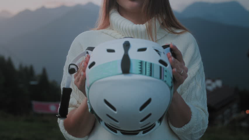 A beautiful Caucasian girl puts a white ski or snowboard helmet on her head. Portrait of a pretty woman at a winter resort. The female wears an orange protective mask or red glasses over her eyes.