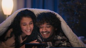 Happy family boyfriend and girlfriend shopping with phone browsing mobile app together smiling excited watching social media video talking couple Indian man and Arabian woman night home under blanket