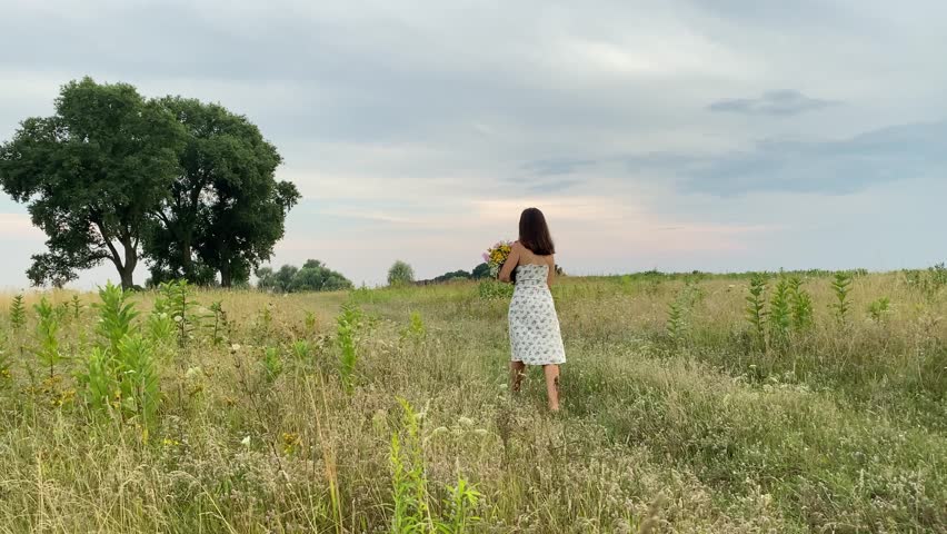 Rear view young adult woman holds bouquet vibrant wildflowers, strolls country road, embraces enchanting rural landscape. Meadow field with cloudy sky at sunset creating mesmerizing countryside scene