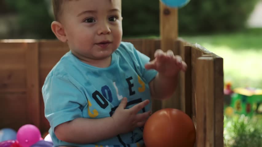 A little boy plays with colorful balls in a wooden enclosure on a grass lawn. Summer, sunny day