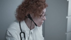 Female senior doctor therapist in white coat and headset talking video conferencing on laptop in office using online video calling application. Remote medical care for patients, online consultation.