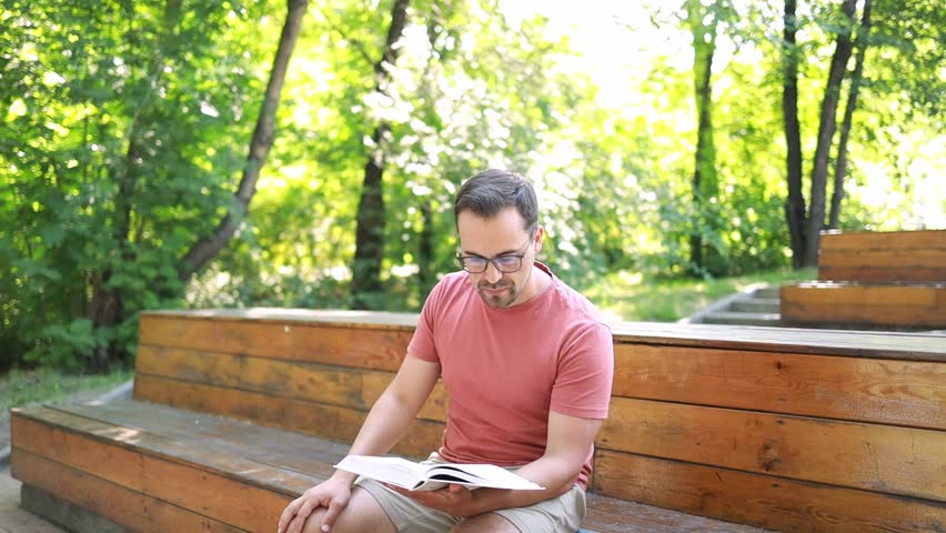 A man reads a book sitting on a park bench on a summer day.