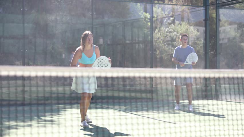 Portrait of emotional fit young woman playing padel tennis on open court in summer, swinging racket to return ball over net. Sportswoman ready to hit volley