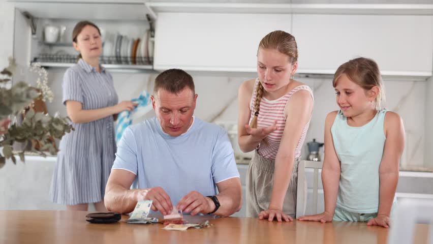 Interested teenage sisters asking for pocket money from smiling father sitting at table in kitchen, counting received salary. Happy woman looking at family while doing household chores in background Royalty-Free Stock Footage #1106824591