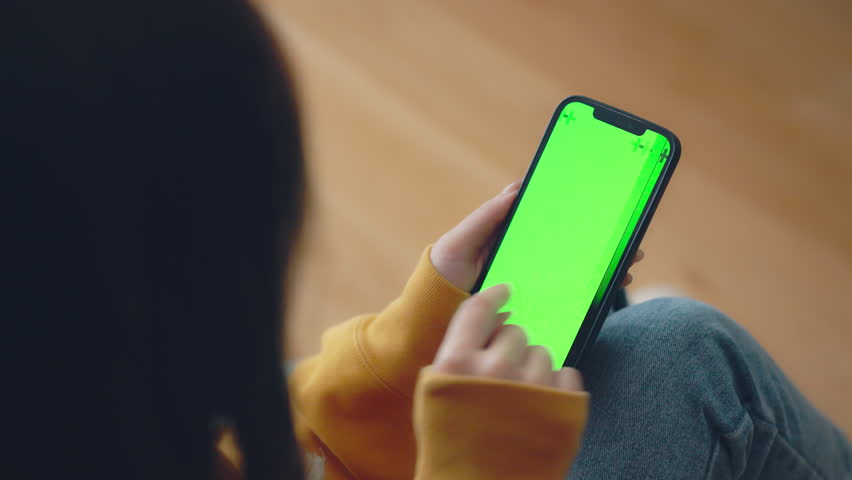 Hand of woman holding mobile smartphone and swipes photos or pictures with blank green screen mock up while sitting on a sofa at home.