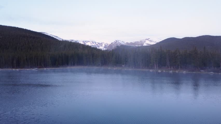Flying over a steaming lake towards the snowcapped Rocky Mountains in Colorado during blue hour, aerial
