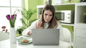 Young beautiful hispanic woman eating cookies using laptop doing video call at dinning room