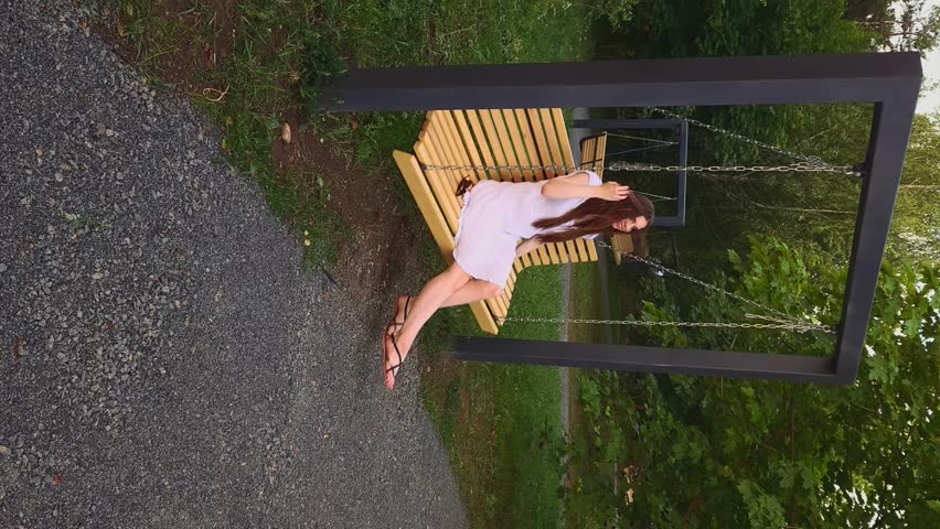 Young woman in white sundress and sandals carelessly swinging on swing in city park