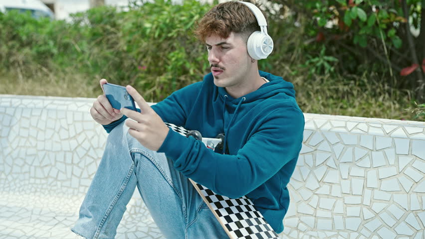 Young hispanic man holding skate playing video game at park