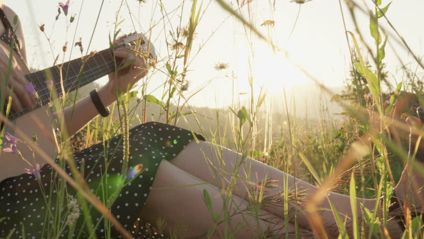 A young woman plays guitar while sitting on the grass amidst wildflowers in summer. A magical atmosphere relaxation, doing your favorite hobby alone in nature, at sunset. Girl makes music and relaxes