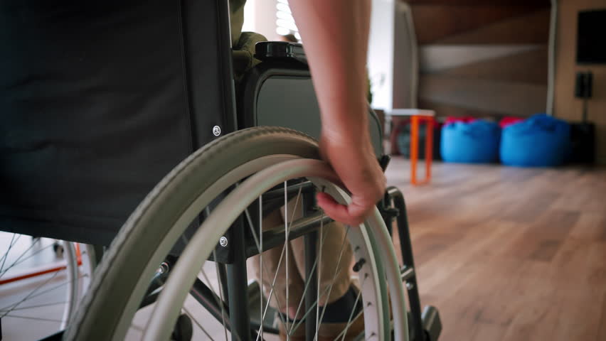 Man with spinal cord injury moves around light room. Male hand turns large wheel of wheelchair. Medical equipment for people with paralysis closeup slow motion