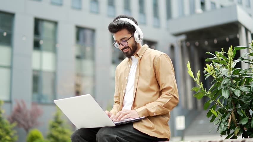 Young employee in headphones works on a laptop while sitting on a bench near the office building. Smiling handsome freelancer is chatting online with a client. IT specialist programmer uses a computer | Shutterstock HD Video #1106849989
