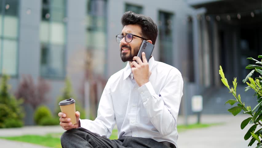 A young businessman talking on a smartphone while sitting on a bench on the street near an office building. Bearded smiling freelancer having a conversation with a colleague or client, drinking coffee