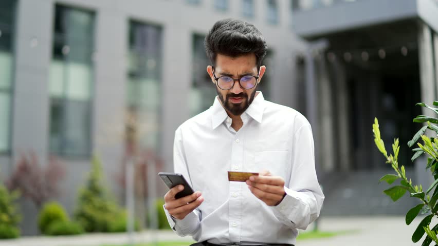 Frustrated stressed man detected fraud while entering credit card number on smartphone while standing on the street near an office building. Worried male is sad that money was stolen from his account Royalty-Free Stock Footage #1106850033