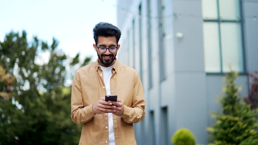 A smiling student is using a smartphone while walking around the campus space near the university building. Happy handsome bearded male checking email, chatting online, browsing social media, texting Royalty-Free Stock Footage #1106850051