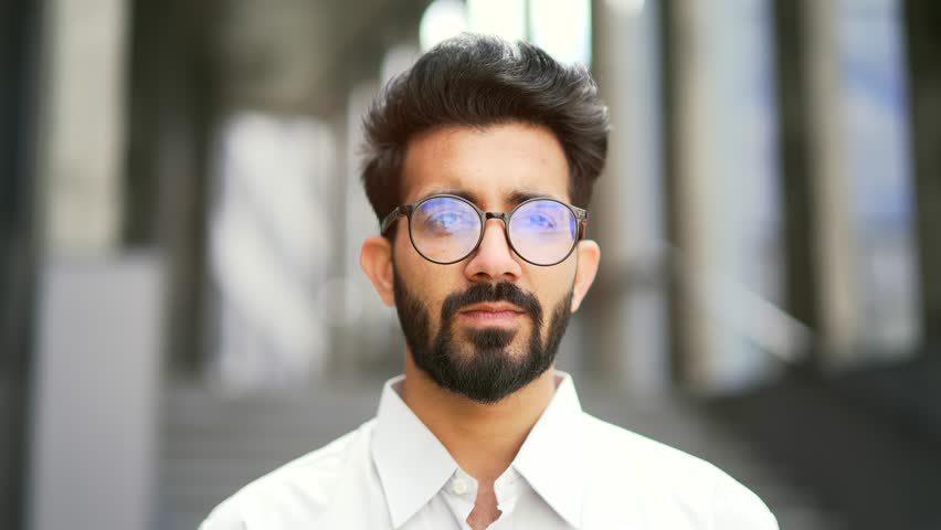 Close up portrait of a confident businessman in a white shirt and glasses looking at the camera. Handsome serious bearded male posing standing on the street in front of an office building. Head shot