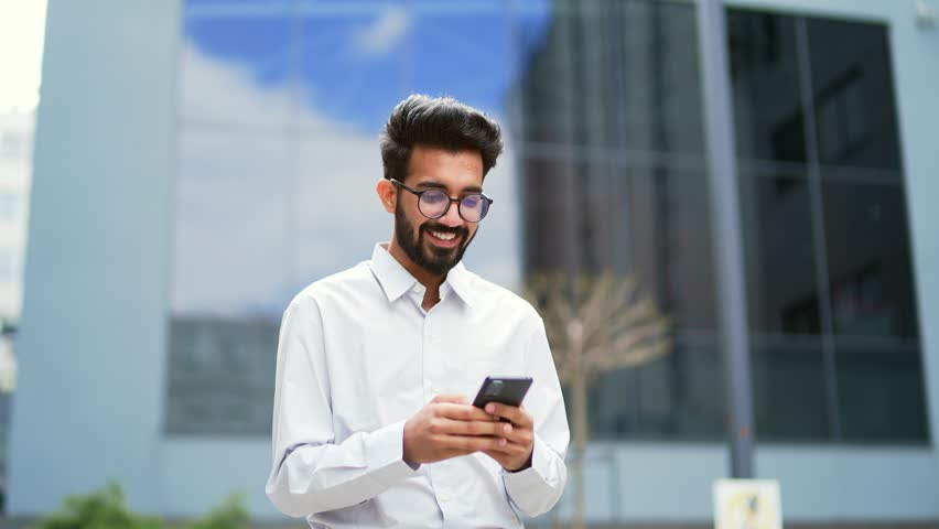 A young smiling businessman is using a smartphone while walking along the street near an office building. Happy handsome bearded male texting, checking email, chatting online, browsing social media Royalty-Free Stock Footage #1106850063