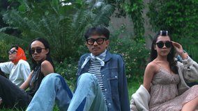 Camera slowly zooms in on a group of diverse, stylish friends relaxing on grass. The focus settles on the young man in the center wearing a denim jacket, striped shirt, neckerchief, and yellow-tinted. - Powered by Shutterstock - Get 15% off with code: PIKWIZARD15