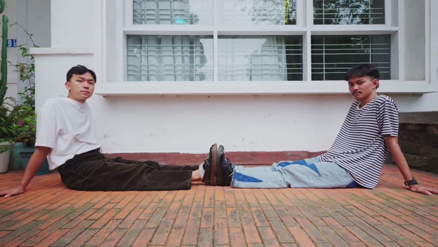 Two young men in retro-inspired fashion sit back-to-back on a brick patio in front of a white building. They lean back on their hands and look directly at the camera with a neutral, cool expression.
