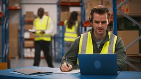 Male worker wearing headset and high vis vest sitting at laptop in busy distribution warehouse - shot in slow motion - Powered by Shutterstock - Get 15% off with code: PIKWIZARD15