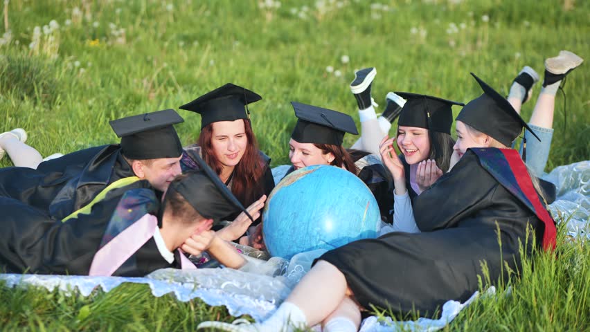 Graduates in black robes looking at a globe lying on the grass. | Shutterstock HD Video #1106852283