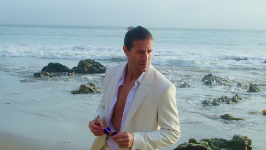 Handsome man in white suit posing by Pacific Ocean. Man holding stylish sunglasses on the beach Royalty-Free Stock Footage #1106857333