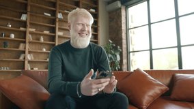 Positive guy using apps playing mobile game chatting in social media surfing web on phone in modern home. Young smiling albino man holding smartphone device texting sms message sitting at home office - Powered by Shutterstock - Get 15% off with code: PIKWIZARD15