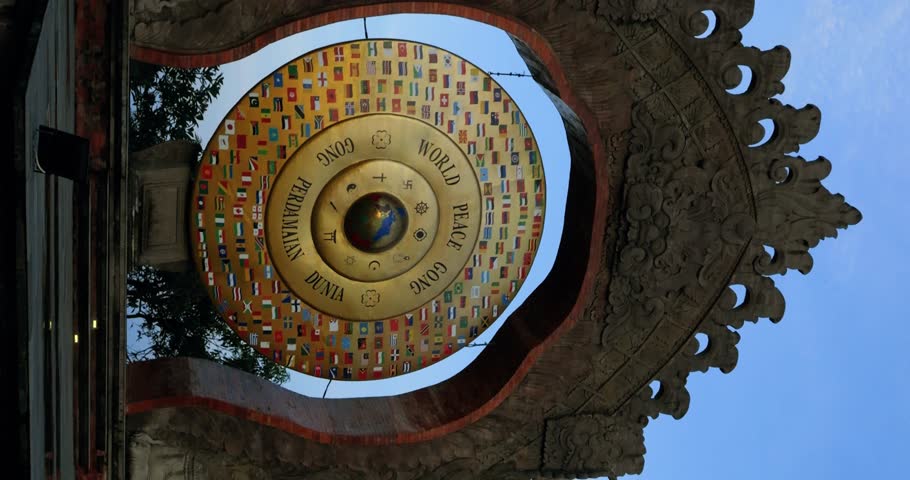 Vertcial Stunning parallax shot of the World Peace Gong in Bali adorned with golden peace signs and national flags, against a backdrop of palm trees, the moon and a vibrant blue sky