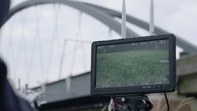 Handheld camera perspective of a white drone with four rotors, flying near a modern cable-arch bridge, with overcast gloomy sky backdrop from monitor perspective