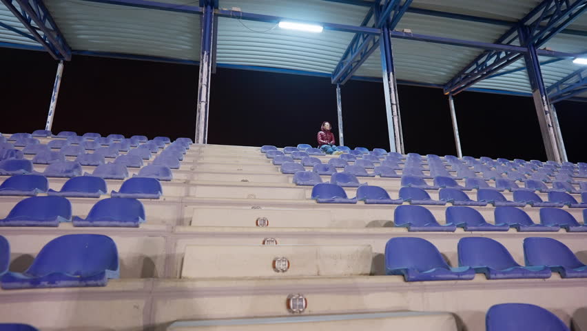 Wide shot stadium tribunes with blurred Caucasian woman sitting on top. Unrecognizable lady enjoying leisure thinking at night on athletic field chair