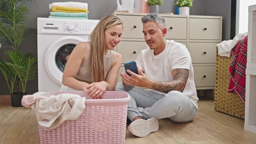 Man and woman couple washing clothes using smartphone at laundry room